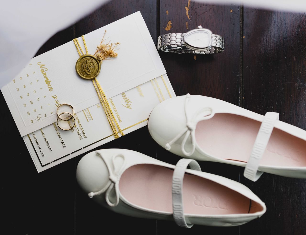 about-img A stylish flat lay of wedding essentials including shoes, invitation, rings, and a watch on dark wood.